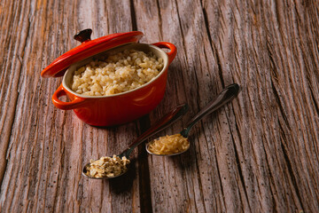 A small red pan of rice, next to two spoons with grains of food, on top of a wooden surface