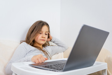 Emotional portrait of attractive cute girl 6-7 old she is tired in a bad mood sitting using laptop on distance learning