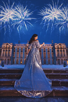 Beautiful girl in a shiny silver dress with a long train standing on the stairs against the background of the palace, the night sky and magnificent fireworks