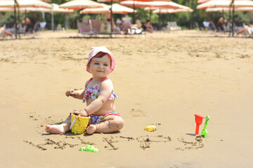 Cute adorable happy baby girl playing on the sandy beach near the sea. Family on resort. Summer holiday concept.