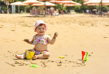 Cute adorable happy baby girl playing on the sandy beach near the sea. Family on resort. Summer holiday concept.