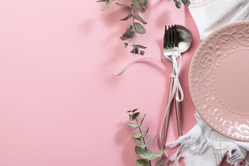 Pink empty plate, white napkin, cutlery set with bow and decorative eucalyptus twigs on pink pastel background. Festive table setting for Valentine’s Day or Easter concept.