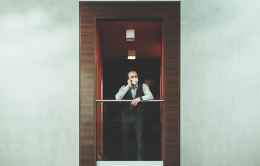 A mature hispanic businessman in an antivirus protective face mask is standing on a balcony in the nook of a modern office chillout area and talking on the phone; copy-space areas on left and right