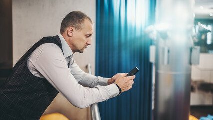 A serious mature businessman is replying to a working correspondence e-mail using his smartphone while leaning against a balcony railing in the office coworking area; a copy space place on the right