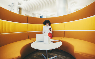 A wide-angle shot of a young elegant African-American businesswoman using her smartphone and gadgets while sitting on a round yellow sofa in office coworking area; woman entrepreneur with a cellphone
