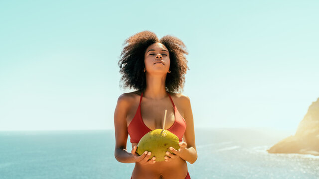 Dazzling young African-American female with curly Afro hair and coconut with a straw in her hands is raising her head up enjoying the sunlight while standing on the cliff of a seaside resort