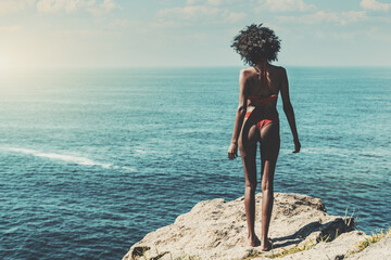 A slim beautiful young black female in swimwear and with curly African hair is standing on a high cliff of a seaside resort, tanning and enjoying observing the endless ocean horizon