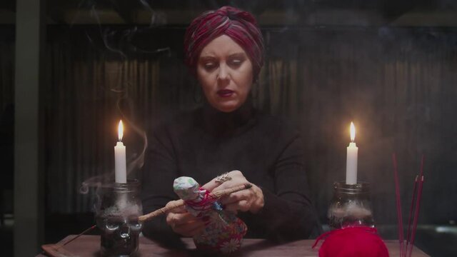 Senior witch woman fortune teller sticks a needle into voodoo doll in terrible magic ritual