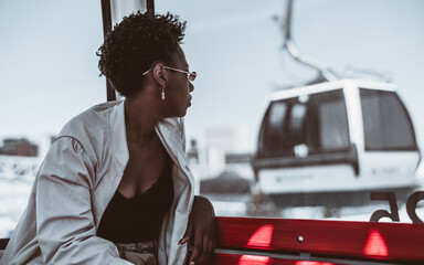 A profile portrait of a young black tourist female hipster in sunglasses and white trench, sitting on a wooden red bench indoors of an overhead cableway car and observing attractions from high above - Powered by Adobe