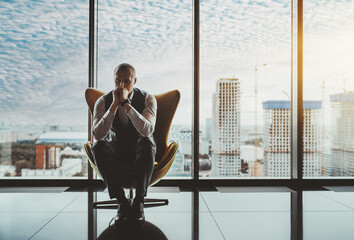 A portrait of a man entrepreneur in a state of worry and dismay sitting on an armchair in front of the window of a luxury office high-rise and thinking about how to solve recent business issues © skyNext