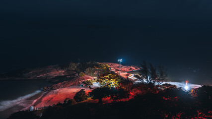 A low-key long exposure shot with a view of Arpoador beach at the night from high above: coastline, floodlight on a mast, dark ocean horizon, blurry trees swaying in the wind, Rio de Janeiro, Brazil