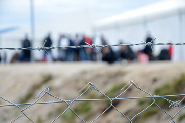 Barbed wire in refugee camp. Migrants behind chain link fence in camp. Group of people behind...