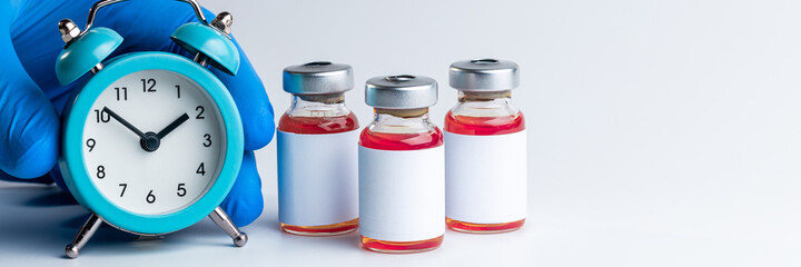medical vaccine vials with red liquid and alarm clock over gray background. time to vaccination...