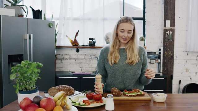 Beautiful young blonde woman uses salt and pepper to prepare a healthy avocado bread for breakfast in modern kitchen