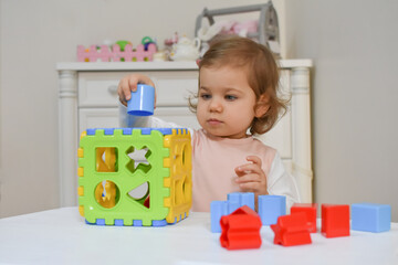 Fototapeta premium Cute adorable baby girl solves sorter puzzle. Toddler Having Fun with educational toys. Kid playing in nursery with nordic stylish. White scandinavian interior for children's room. 