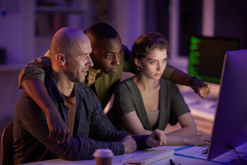 Group of three modern people wearing casual outfits spending evening together working in office looking at computer screen