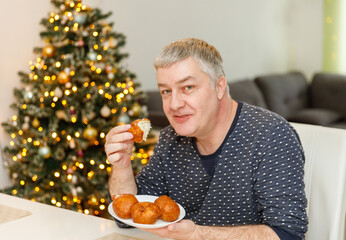 Caucasian man eating olibol near the Christmas tree at home, portret,Oliebollen - Dutch traditional Dessert - Also known as Dutch Doughnuts or dutchies.