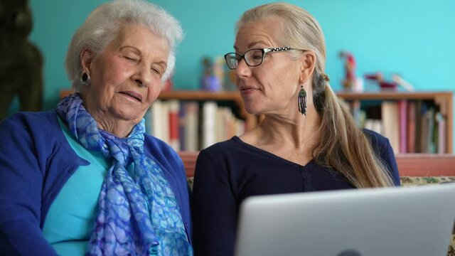 Extreme closeup front view of smiling elderly mother and mature woman sitting on the sofa and shopping online on a laptop computer.