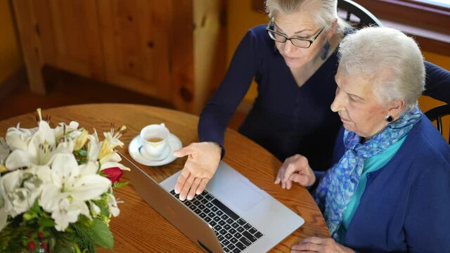 Overhead shot of mature woman and elderly mother working on laptop computer on dining room table having tea.