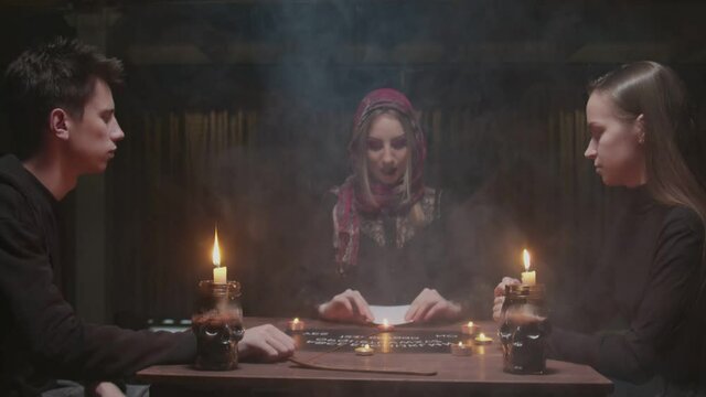 Young couple visits young witch female fortune teller. Young woman shows photo of her lost friend to the witch, then they takes each others hands and closes eyes