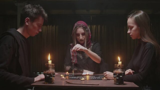Witch female fortune teller tries to speak with ghost. She uses a blue crystal and board with letters. Two women and one man uses Ouija Board Spirit Game