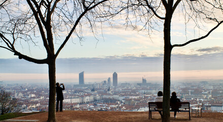 Panoramic picture of 2 people sitting on a bench admiring a wonderful sunrise over the city of Lyon in France and a standing tourist taking pictures