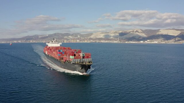 Aerial view of large cargo ship with red containers floating on sea waves on warm sunny day. Concept of work on sea delivery of goods and cargo