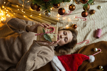 Fototapeta premium Christmas composition. Woman wrapping gift box in craft paper near fir tree, next lying santa hat, candy canes. new year concept. Xmas celebration 2021. Flat lay, top view, overhead