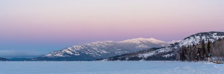 The sun rising over a huge snow capped mountain on a crisp winter morning with clear, bright sky, pink, blue mountains peak with frozen lake and woods in foreground. Pastel skies. 