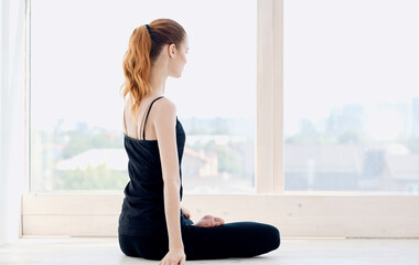 A woman in dark sportswear practices yoga while sitting near the window indoors