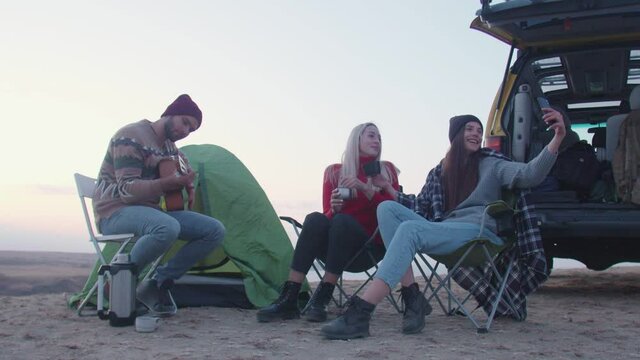Young man plays guitar and two woman sits on campers chairs, drinks a hot tea and takes selfie