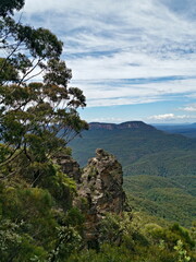 Fototapeta premium Beautiful view of tall mountain peak, with mountains, valleys, trees, blue sky and white clouds in the background ,Three Sisters Lookout, Blue Mountain National Park, New South Wales, Australia 