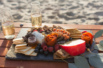 Gazing board of charcuterie. berries, crackers, and cheeses set on wood table with two glasses of champagne with blurry beach sand in the background.