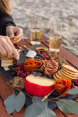 Fototapeta na wymiar Woman's hand cutting into soft goat cheese onto a cracker from a grazing board of charcuterie on a wood table with two glasses of champagne with blurry beach sand in the background.
