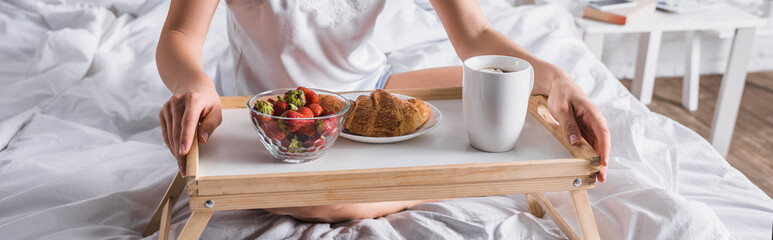 cropped view of woman having croissant and strawberry for breakfast in bed, banner