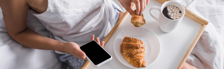 cropped of woman having croissant and cocoa for breakfast while using smartphone in bed, banner