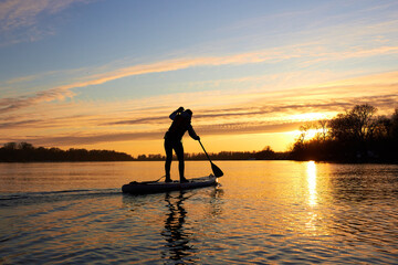Silhouette of woman paddle on stand up paddle boarding (SUP) on quiet winter river at sunset