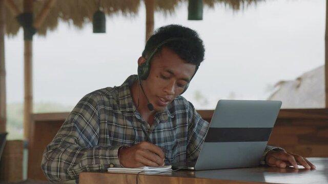 Lockdown of young Asian male helpdesk agent wearing checkered shirt and headset sitting at table on terrace in front of laptop, communicating with client by phone and writing in notebookLockdown of yo