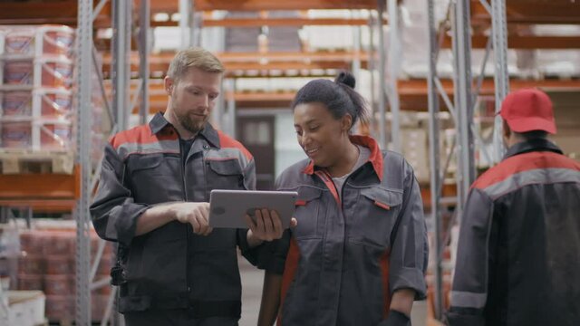 Medium front-view footage of couple of multi-ethnic man and woman in blue overalls walking towards camera along tall warehouse discussing construction project looking at digital tablet