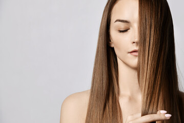Portrait of sensual young woman with closed eyes and naked shoulders holding strand of her long silky hair covering half of her face over grey background with copy space. Haircare, beauty, wellness