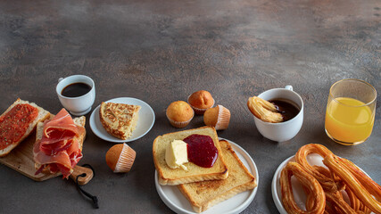 Spanish breakfast assortment. Tortilla, bruschetta with jamon and tomatoes,  crusty toast with butter and jam, muffins, coffee and orange juice on concrete background.. Large image for banner.