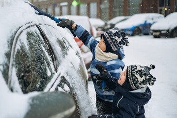 cute caucasian boy wearing scandinavian knit hat brushing snow from a car on winter day  near...