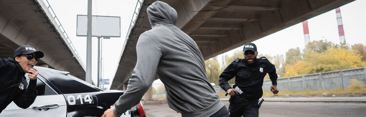 hooded offender running from multicultural police officers near patrol car on urban street, banner. © LIGHTFIELD STUDIOS