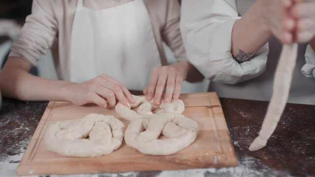 Midsection footage of little girl and woman in cooking aprons shaping homemade pretzels on wooden cooking board at kitchen table