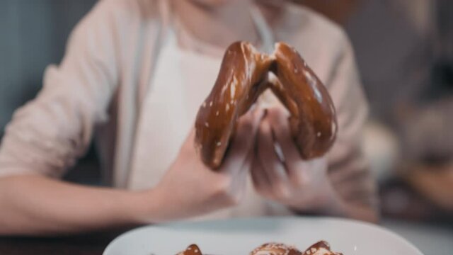 Tilting-up medium close-up of pretty young girl holding freshly baked pretzel splitting it to share with mom standing at cooking table