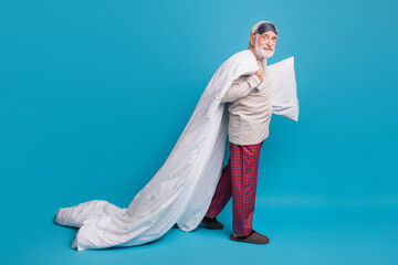 Full length profile side photo portrait of old man in eye mask walking with pillow blanket isolated on pastel blue colored background