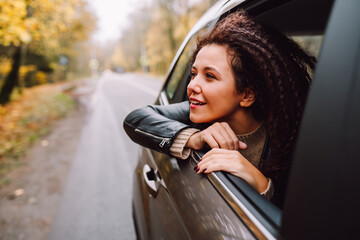 Fototapeta na wymiar Young afro hair woman travel by car on wild forest autumn road. Female look in open window from back sit with happy smile.
