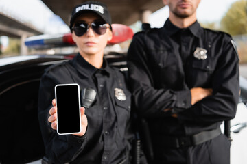 Smartphone with blank screen in hand of policewoman near colleague and car on blurred background.