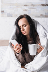 sick young woman with hot drink and tissue using smartphone in bed
