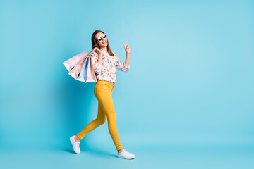 Profile photo of young lady go carry shop bags show v-sign wear sunglass geometry print shirt yellow pants isolated blue color background